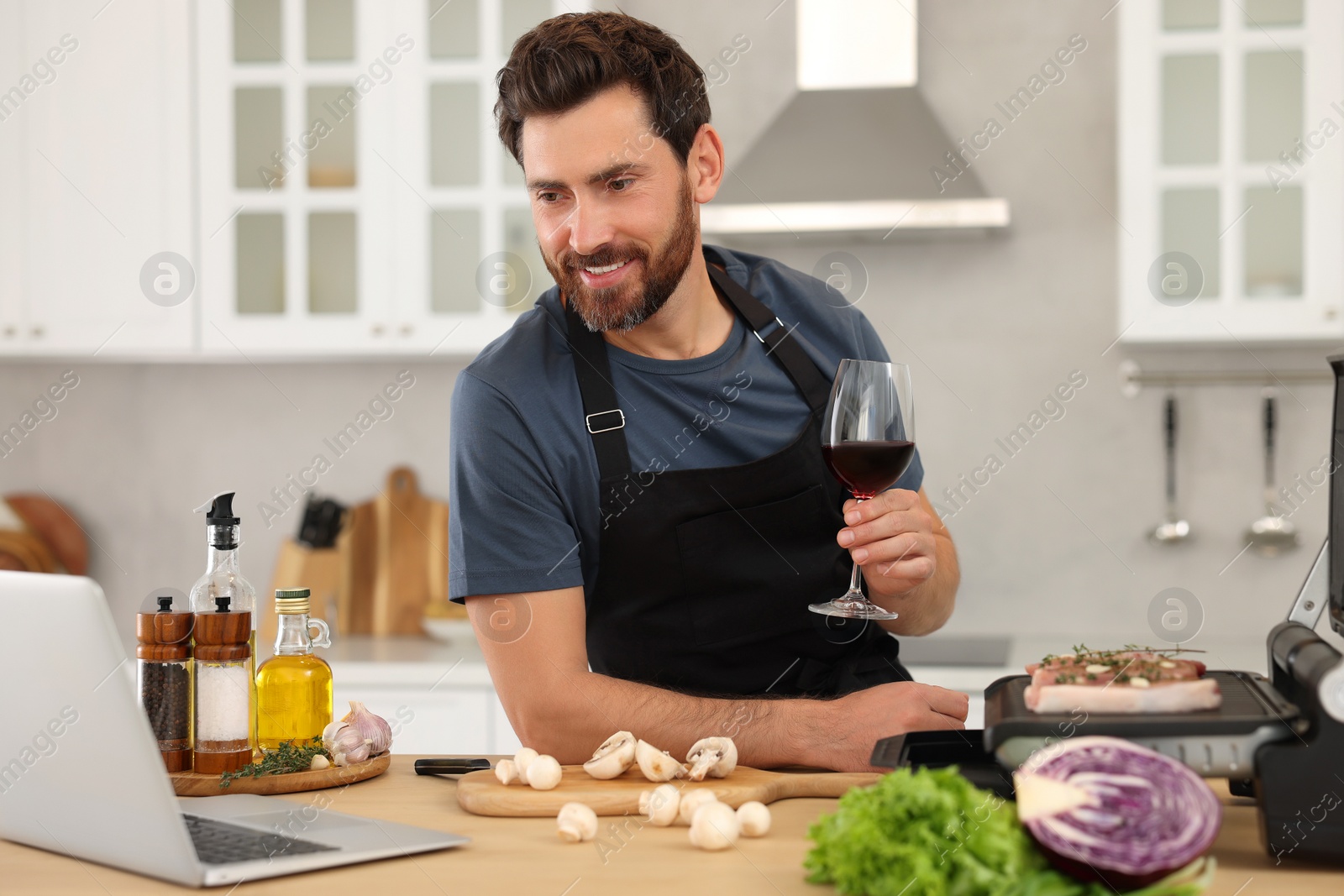 Photo of Man with glass of wine making dinner while watching online cooking course via laptop in kitchen