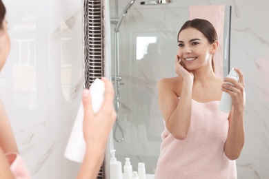 Photo of Young woman applying thermal water on face near mirror in bathroom. Cosmetic product