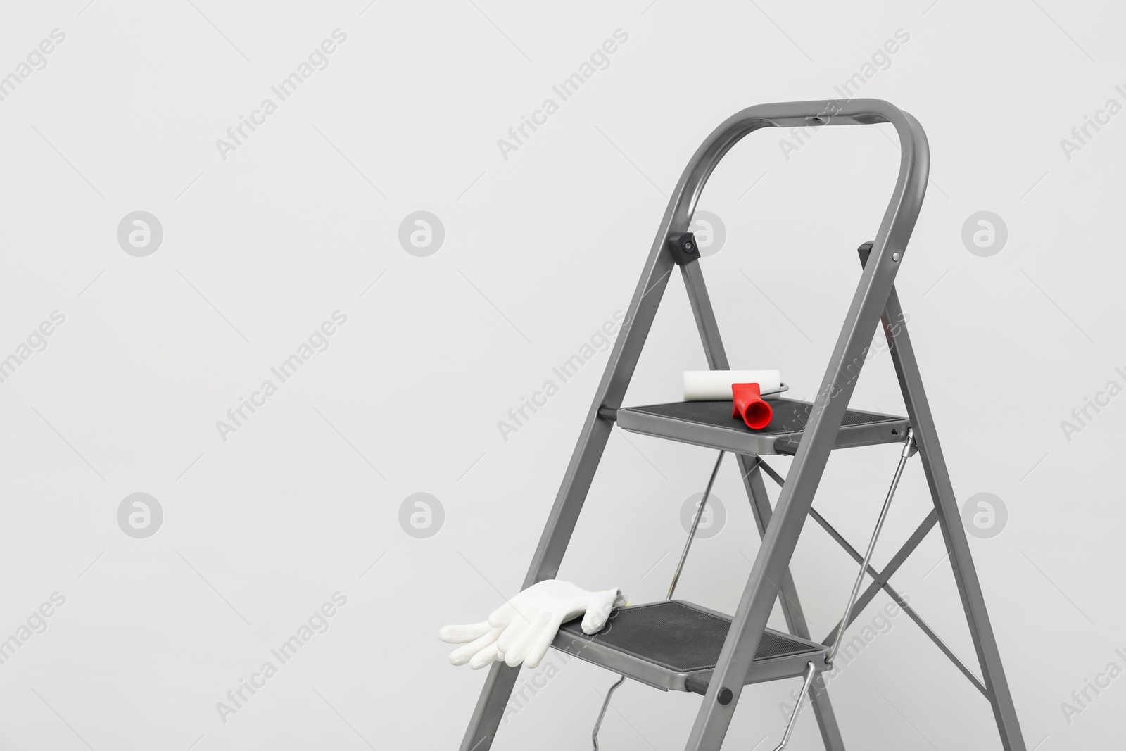 Photo of Metallic folding ladder, paint roller brush and rubber gloves near light grey wall, space for text