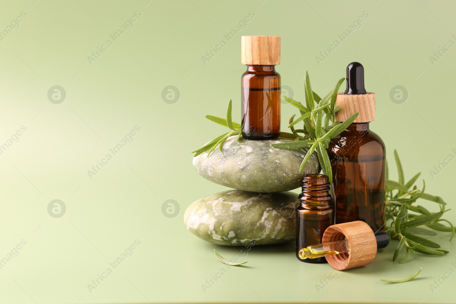 Photo of Aromatic essential oils in bottles, rosemary, pipette and pebble stones on green background. Space for text