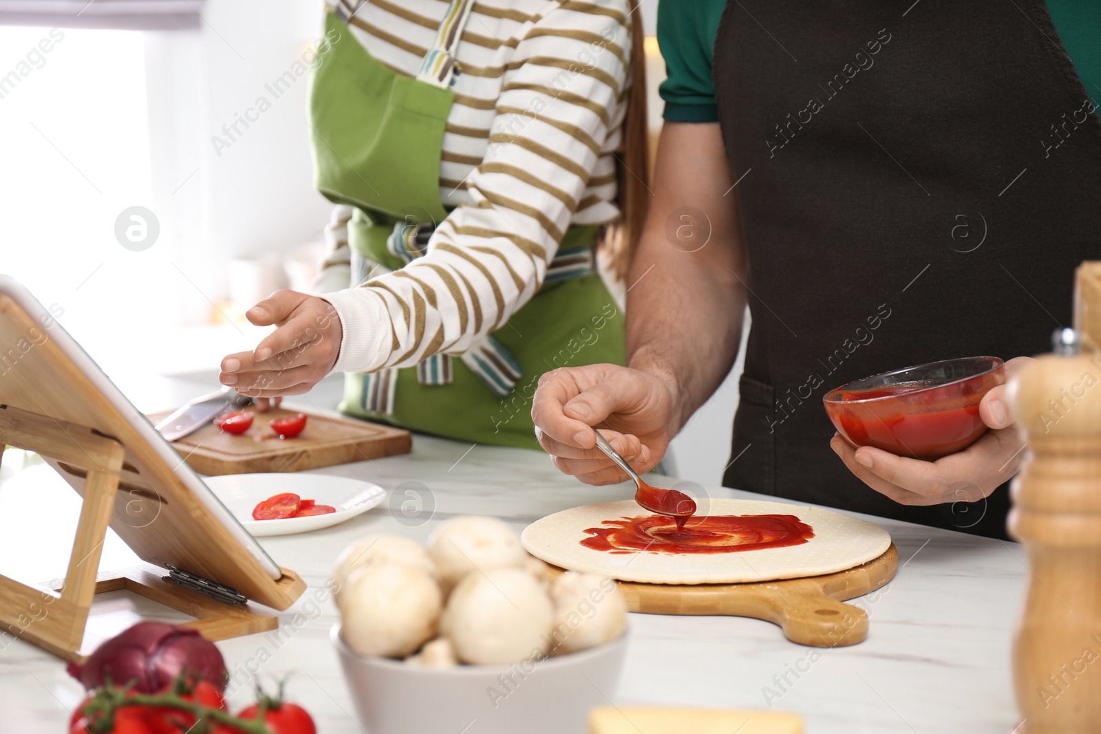 Photo of Couple making pizza together while watching online cooking course via tablet in kitchen, closeup