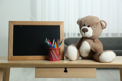 Photo of Teddy bear, small blackboard, pencils and pens in holder on wooden table indoors. Space for text