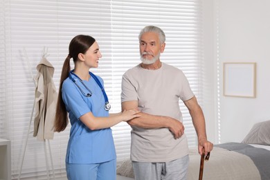 Young healthcare worker assisting senior man with walking cane indoors