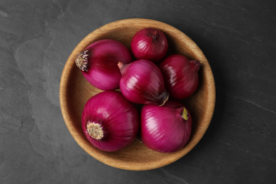 Ripe red onion bulbs in wooden bowl on black table, top view