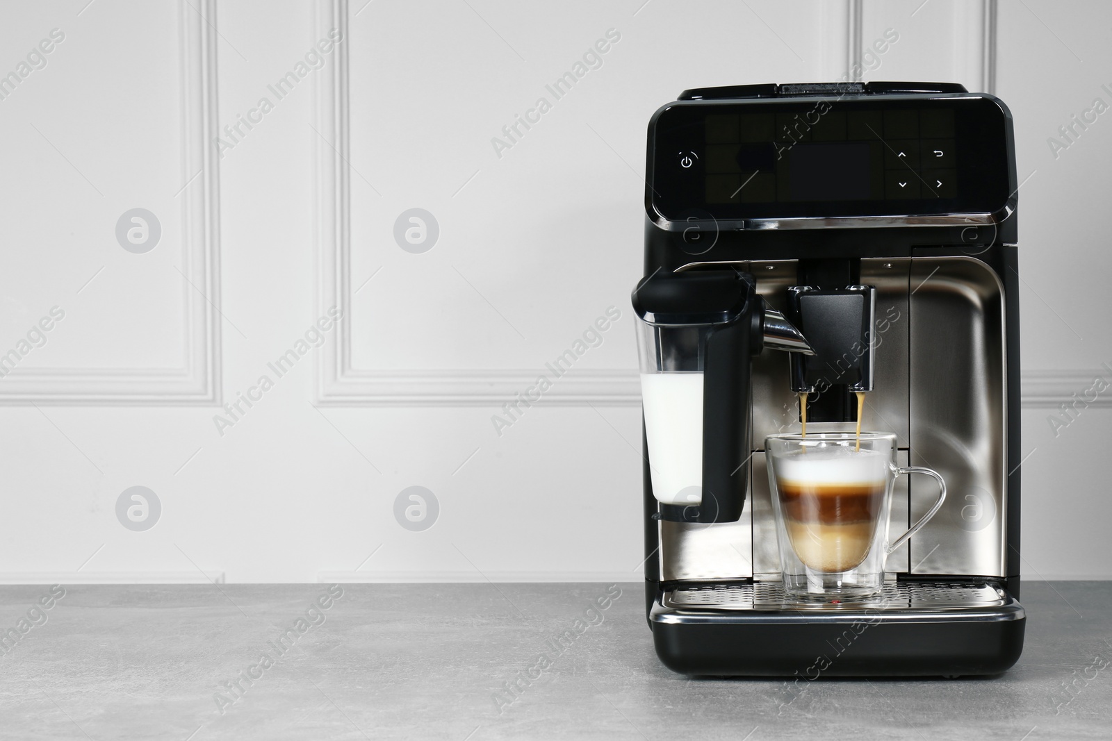 Photo of Modern coffee machine making latte into glass cup on grey table near white wall. Space for text