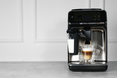 Photo of Modern coffee machine making latte into glass cup on grey table near white wall. Space for text