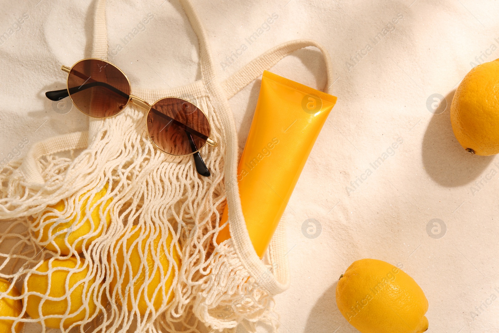 Photo of String bag with sunglasses, lemons and sunscreen on sand, flat lay