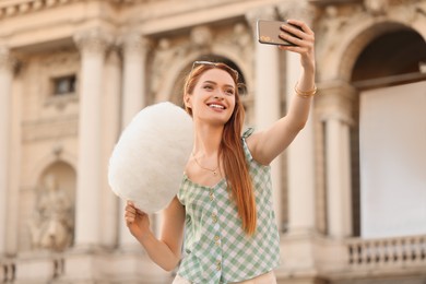 Photo of Smiling woman with cotton candy taking selfie on city street