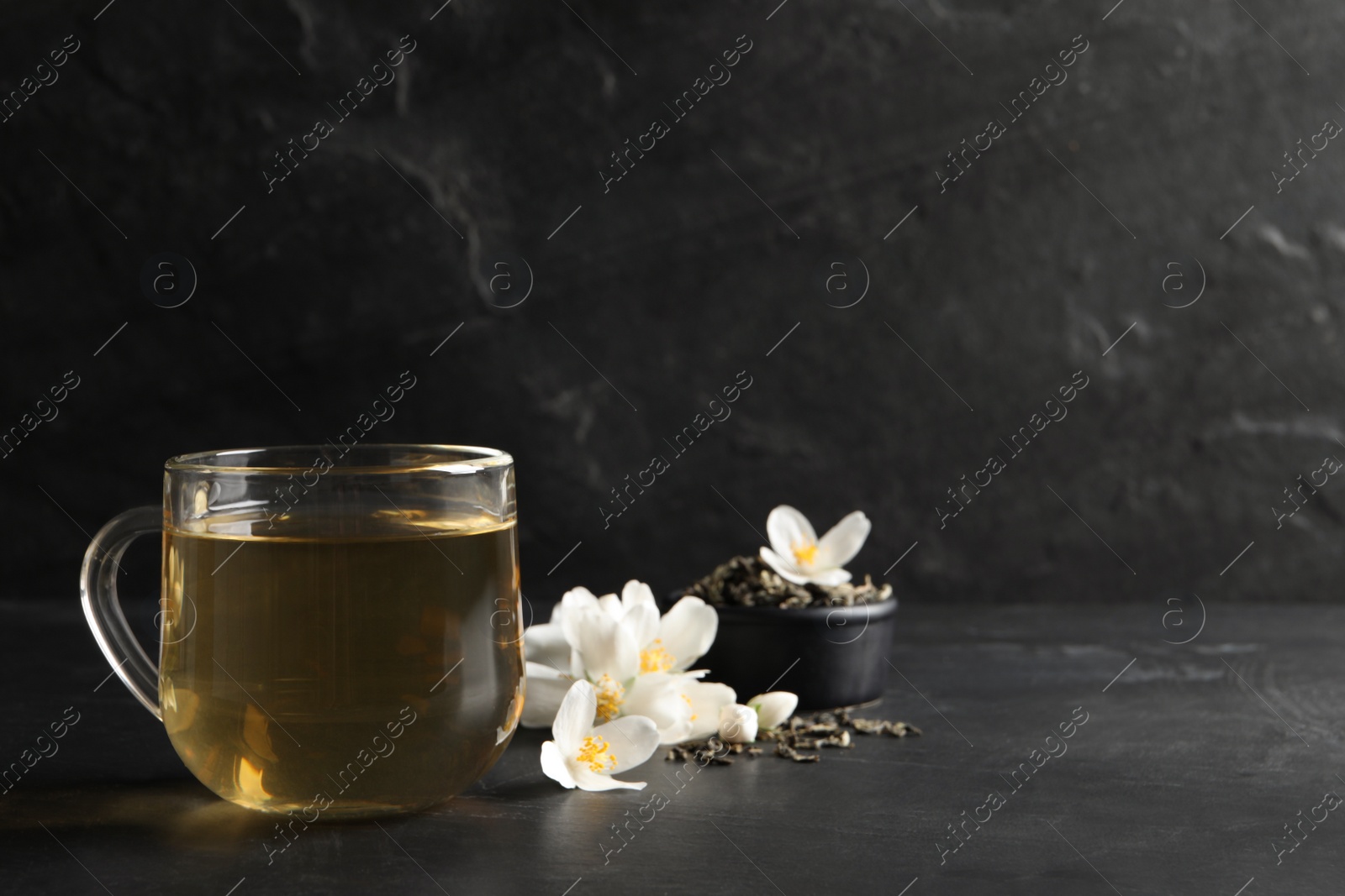 Photo of Glass cup of jasmine tea and fresh flowers on black table. Space for text