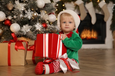 Photo of Cute baby in Santa's elf clothes holding gift box near Christmas tree at home