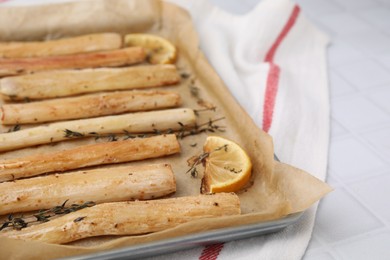 Photo of Baking tray with cooked salsify roots, lemon and thyme on white tiled table, closeup