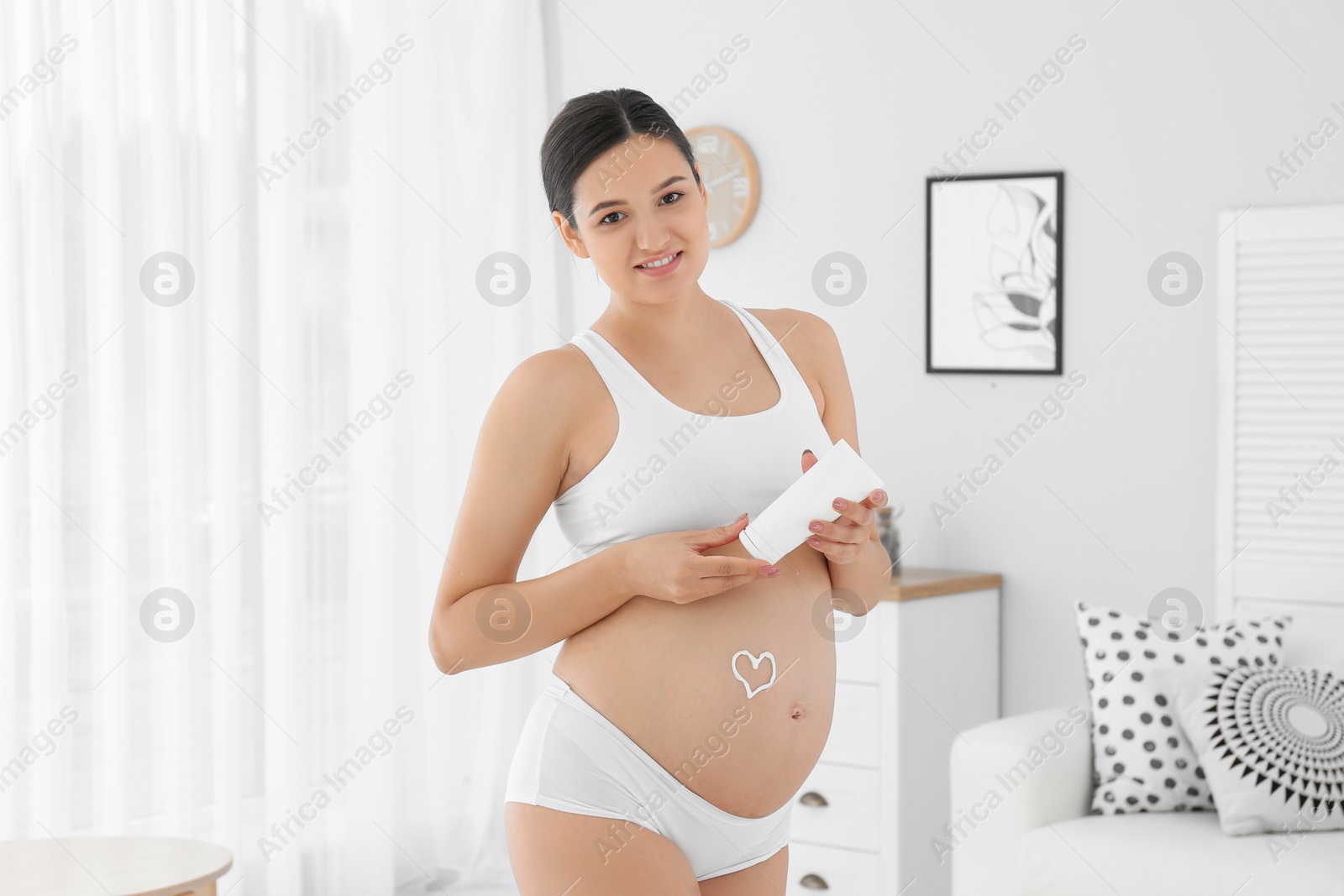 Photo of Pregnant woman with heart on her belly holding body cream at home