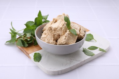 Photo of Bowl with pieces of tasty halva and mint leaves on white tiled table