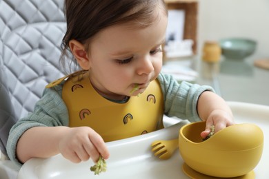 Cute little baby eating healthy food in high chair indoors, closeup