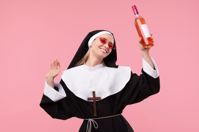 Photo of Happy woman in nun habit and sunglasses holding bottle of wine on pink background