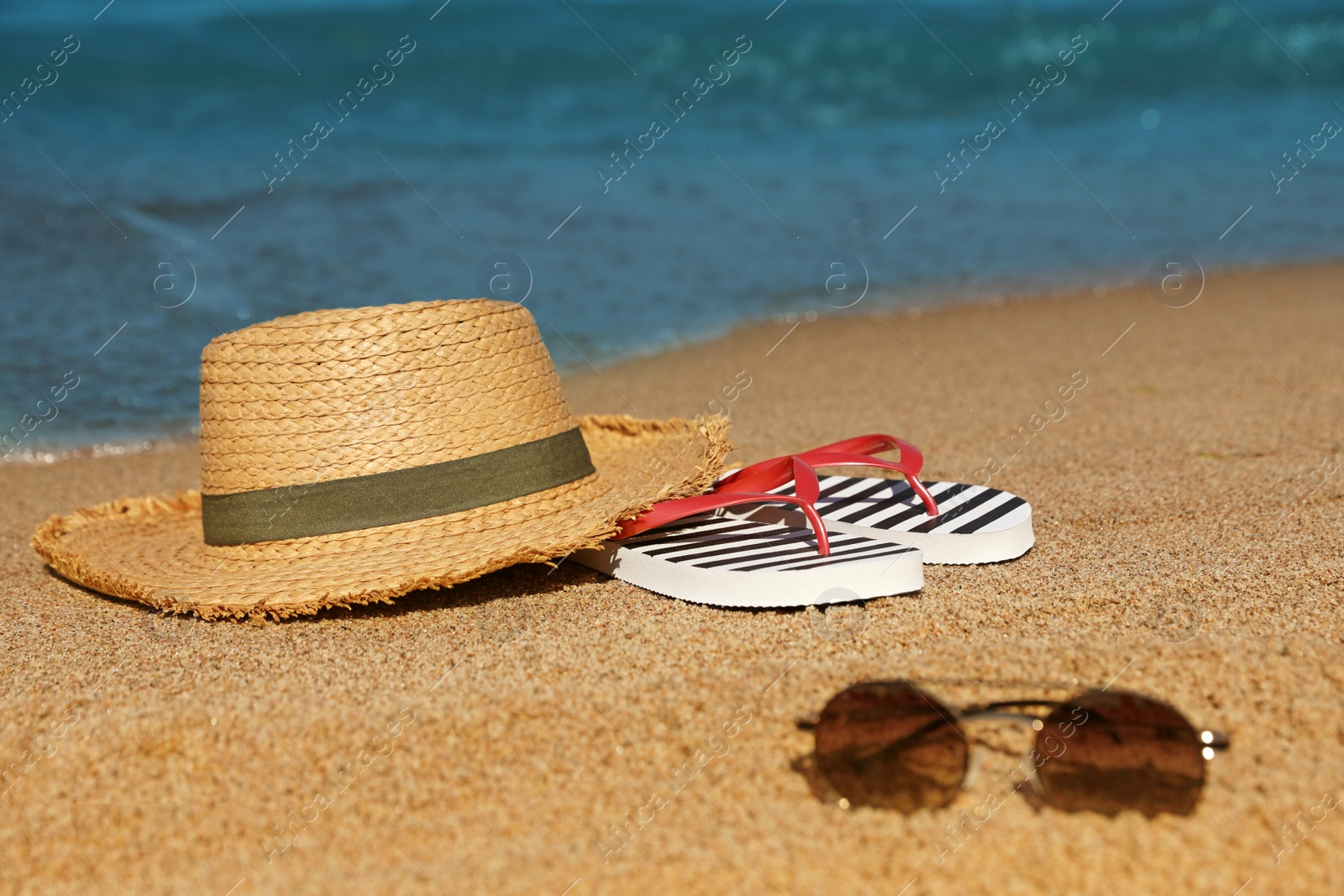 Photo of Stylish straw hat, sunglasses and striped flip flops on sandy beach near sea