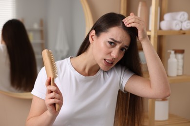 Photo of Emotional woman with brush examining her hair in bathroom. Dandruff problem