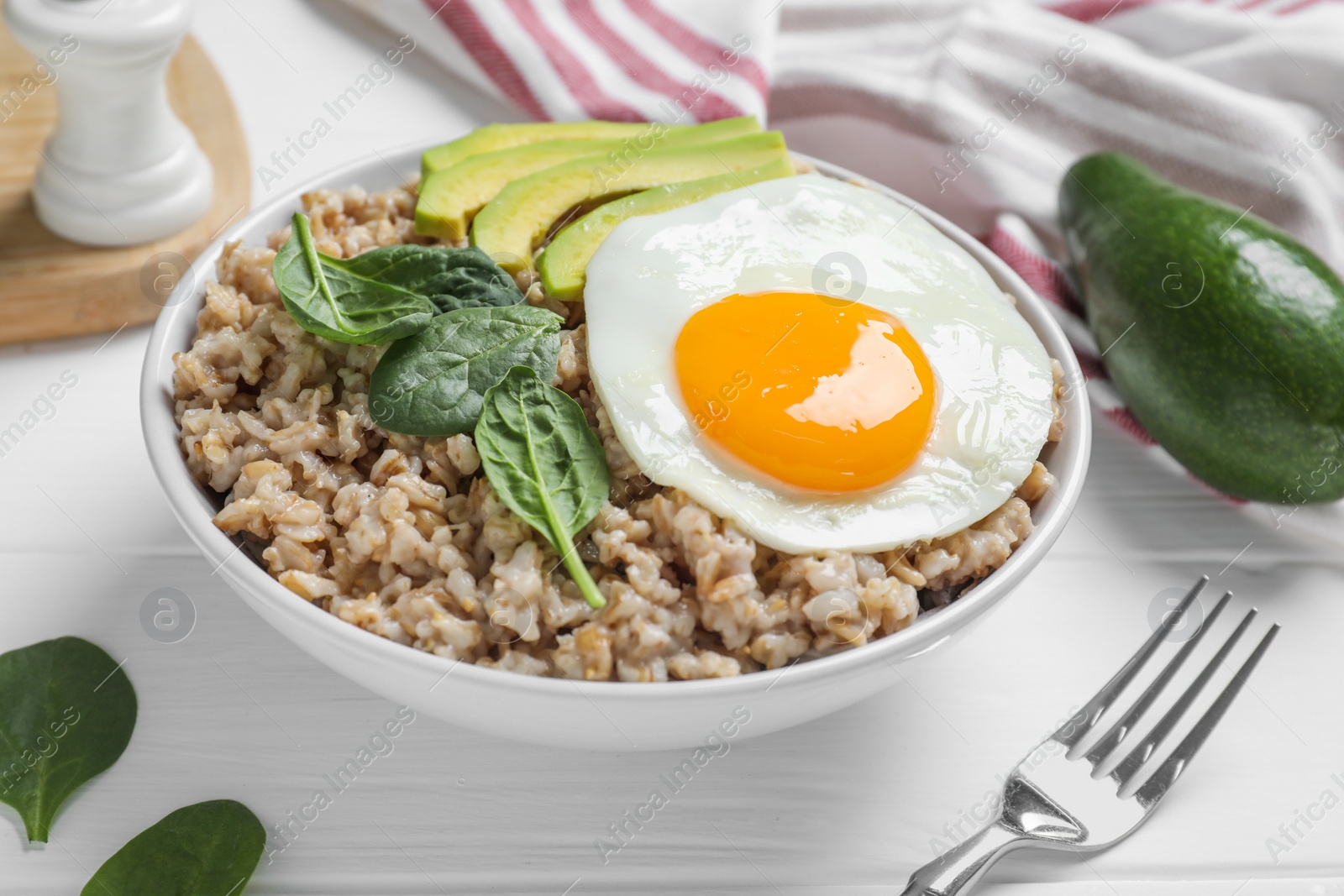 Photo of Tasty boiled oatmeal with fried egg, avocado and basil served on white wooden table, closeup