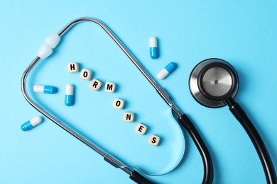 Photo of Word Hormones made of wooden cubes with letters, stethoscope and pills on light blue background, flat lay