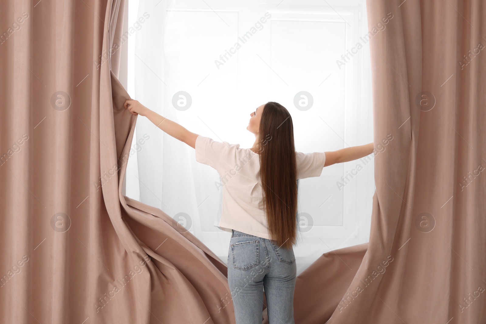 Photo of Woman opening stylish curtains at home, back view