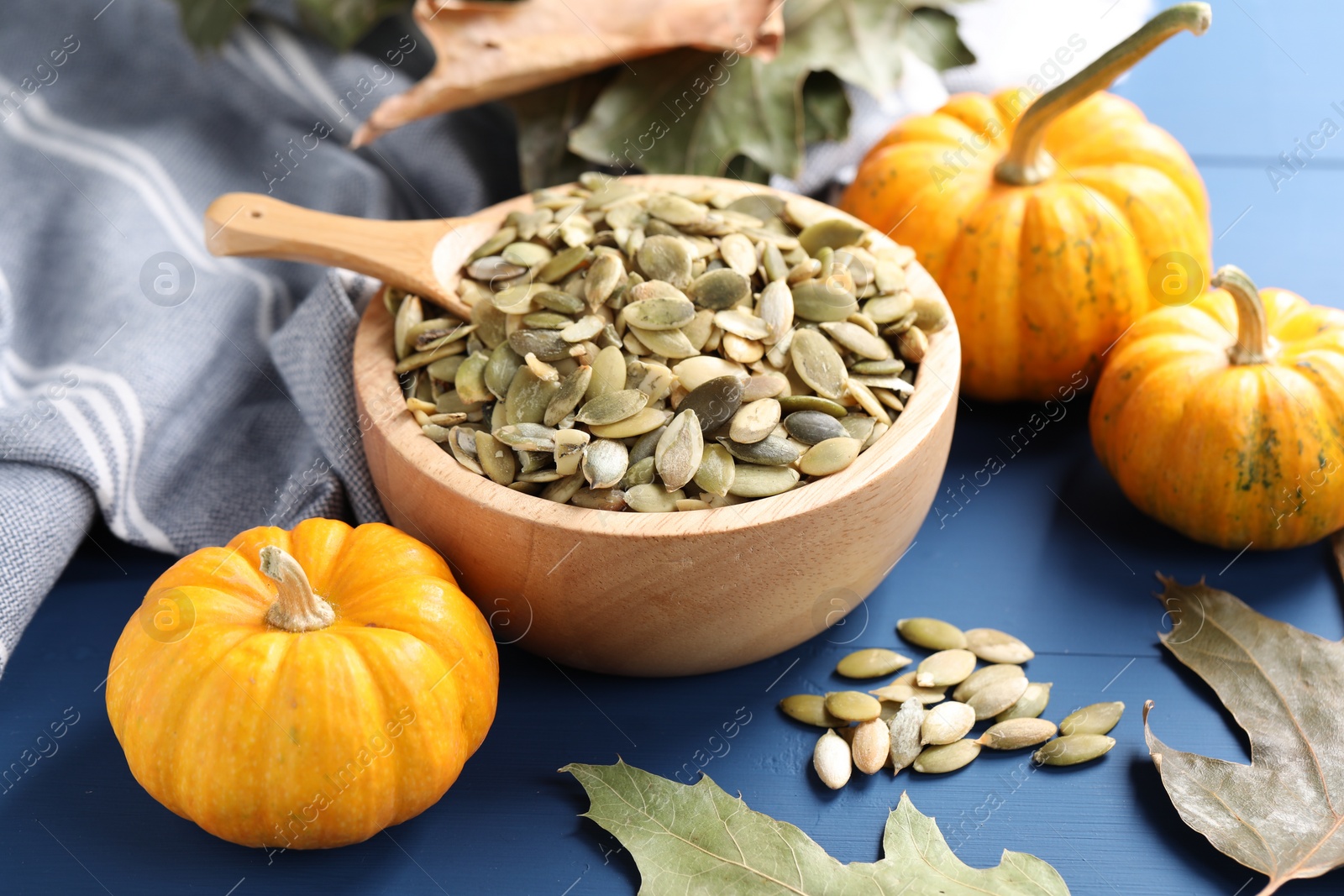 Photo of Bowl with seeds, fresh pumpkins and dry leaves on blue wooden table