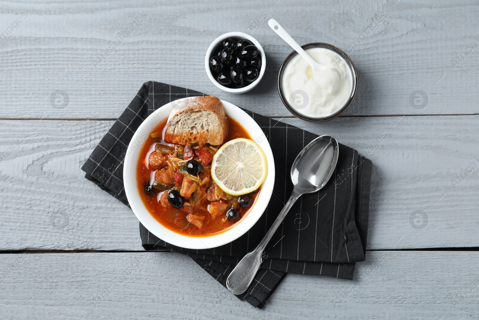 Photo of Meat solyanka soup with sausages, olives and vegetables served on grey wooden table, flat lay