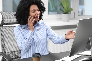 Photo of Young woman talking on phone while working with computer at table in office