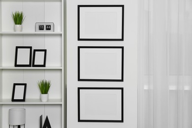 Photo of Stylish room interior with empty frames hanging on white wall near shelving unit