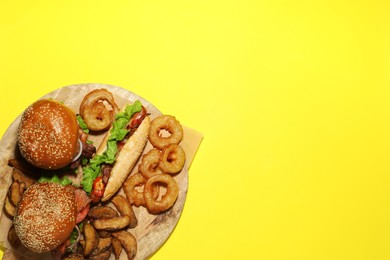 Tasty burgers, hot dog, potato wedges and fried onion rings on yellow background, top view with space for text. Fast food