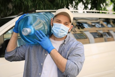 Photo of Courier in medical mask holding bottle of cooler water near car outdoors. Delivery during coronavirus quarantine