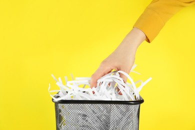 Woman putting shredded paper strips into trash bin on yellow background, closeup
