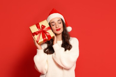 Beautiful young woman in Santa hat with Christmas gift on red background