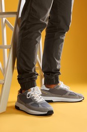 Photo of Man wearing stylish sneakers near white ladder on yellow background, closeup