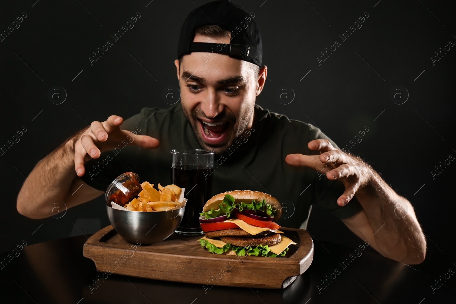Photo of Young hungry man and tasty burger served on wooden board with French fries against black background