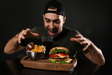 Photo of Young hungry man and tasty burger served on wooden board with French fries against black background