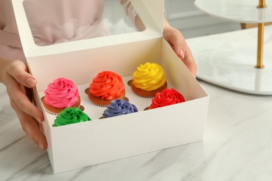 Photo of Woman with box of delicious colorful cupcakes at white marble table indoors, closeup
