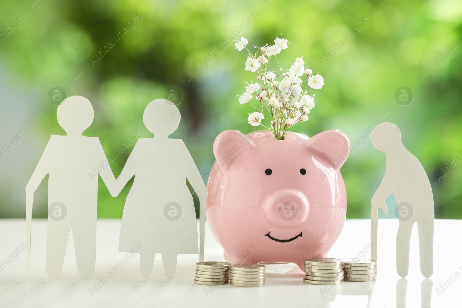 Photo of Pension savings. Figures of elderly people, piggy bank with flowers and coins on white table against blurred green background