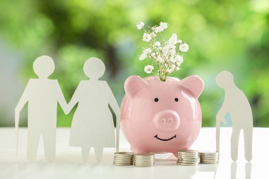 Pension savings. Figures of elderly people, piggy bank with flowers and coins on white table against blurred green background