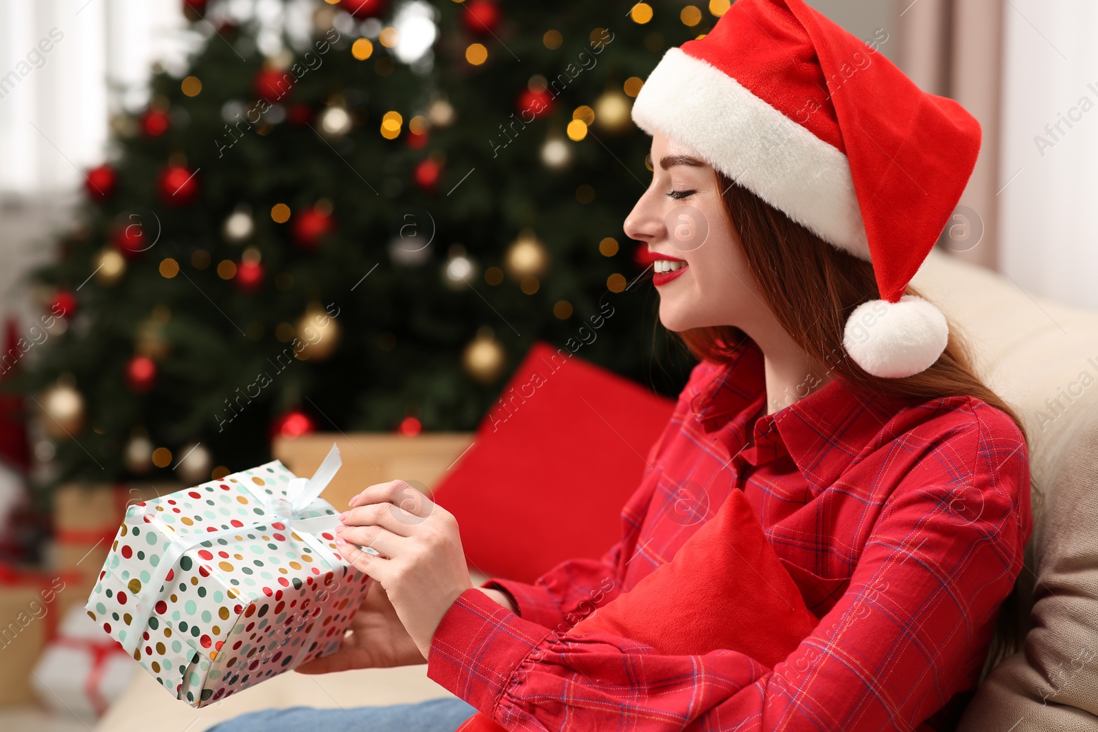 Photo of Beautiful young woman in Santa hat opening gift box near Christmas tree at home