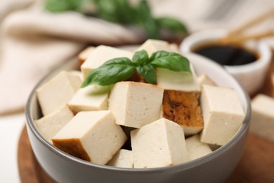 Photo of Bowl of smoked tofu cubes, soy sauce and basil on wooden tray, closeup