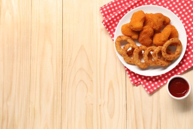 Tasty fried onion rings, chicken nuggets and ketchup on wooden table, flat lay. Space for text