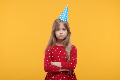 Photo of Unhappy little girl in party hat with crossed arms on yellow background