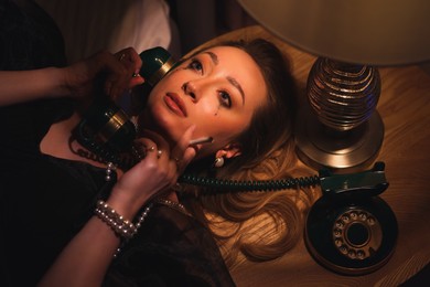 Photo of Young woman with cigarette and vintage telephone crying under dim lamp light indoors in evening