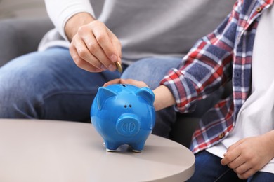 Photo of Little boy with his father putting coin into piggy bank indoors, closeup