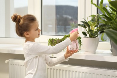 Cute little girl spraying beautiful green plant on windowsill at home. House decor