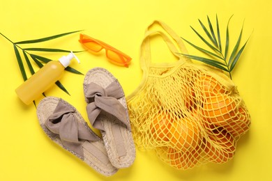 Photo of String bag with oranges, sunglasses, cosmetic product, slippers and palm leaves on yellow background, flat lay