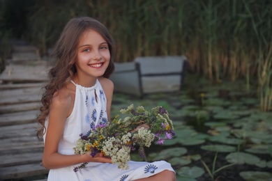 Photo of Cute little girl holding wreath made of beautiful flowers on pier near pond in evening