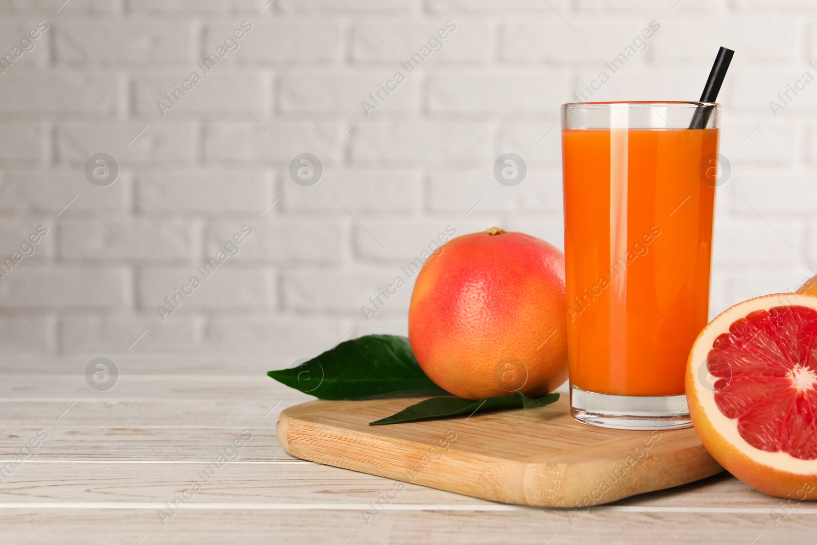 Photo of Tasty grapefruit juice in glass and fresh fruits on light wooden table against white brick wall. Space for text