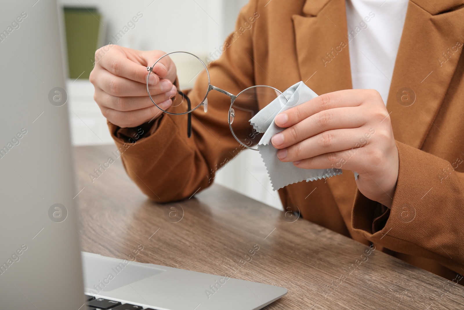 Photo of Woman wiping her glasses with microfiber cloth at wooden table indoors, closeup