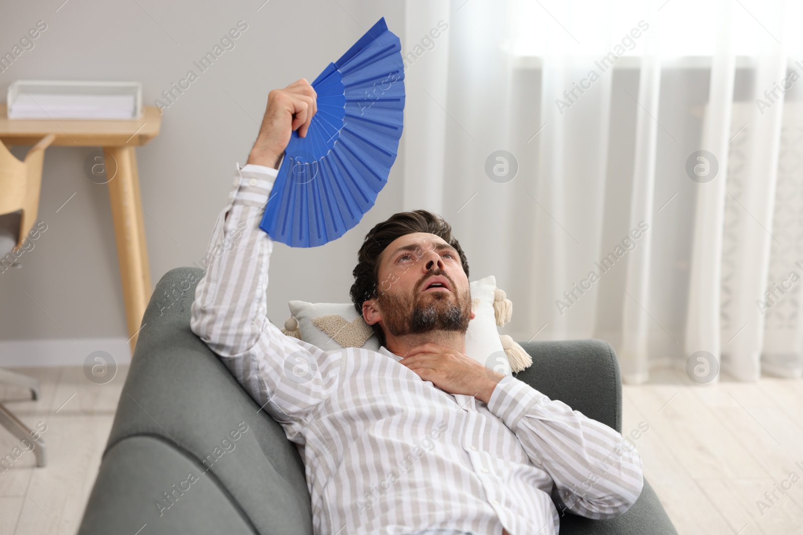 Photo of Bearded man waving blue hand fan to cool himself on sofa at home